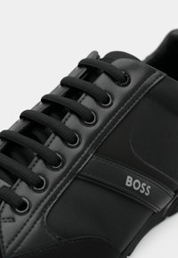 Zwarte sneaker met een gestructureerde afwerking, voorzien van zwarte veters, een glanzend logo accent en een glad paneel aan de zijkant.