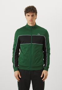 Groene en zwarte zip-up trainingsjas met witte accenten en zijstrepen, voorzien van een hoge kraag en logo op de borst. Zachte stoftextuur.