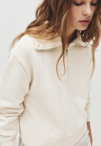 Bež pleteni pulover s ovratnikom na četrt zadrgo, teksturiranim šivom in rebrastim robom. Mehak material s sproščenim krojem.