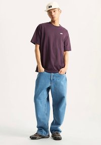 Lila Baumwoll-T-Shirt mit kleinem Logo auf der Brust, kombiniert mit lockeren hellblauen Jeans und dunkel texturierten Schuhen. Akzent durch weiße Kappe.