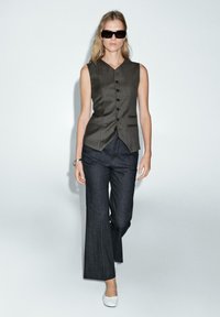 Bezramienna kamizelka w ciemnoszarym kolorze z guzikami z przodu, zestawiona z szerokimi spodniami dżinsowymi w ciemnym kolorze i białymi płaskimi butami, tworzy zorganizowany wygląd.