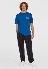 Blue cotton t-shirt with a crew neck and short sleeves, featuring a white logo on the chest. Paired with black cargo pants and white sneakers.