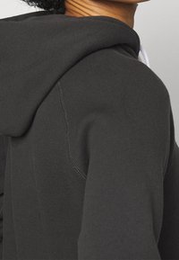 Nahaufnahme einer Person, die einen dunkelgrauen Hoodie trägt, mit sichtbaren Nahtstichen und einer Kapuze, die auf dem oberen Rücken ruht.