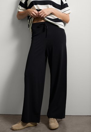 Persona che indossa pantaloni larghi neri con coulisse, sneakers beige e una maglietta bianca con spesse righe orizzontali nere, in piedi contro un muro.