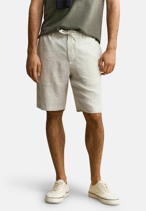 Man in beige linnen shorts met een trekkoord in de taille, een olijfgroen T-shirt en gebroken witte casual sneakers op een witte achtergrond.