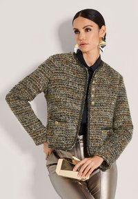 Frau trägt eine strukturierte, mehrfarbige Jacke, metallische Hose, große gold- und silberfarbene geometrische Ohrringe und hält eine reflektierende goldene Clutch. 