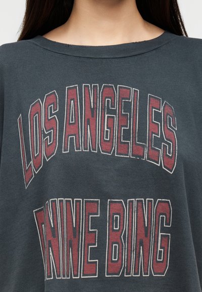 Közeli kép egy sötétszürke ingről, amelyen fakó piros és fehér, nagy betűs felirat olvasható: "LOS ANGELES" és "NINE BING".