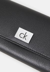 Portefeuille en cuir noir texturé avec une plaque logo rectangulaire en argent portant le motif "ck" en relief. Design compact avec une finition lisse.