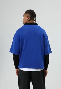 Blauw oversized t-shirt met een zwarte kraag, korte mouwen en een losse pasvorm, gecombineerd met zwarte broek, gezien van achteren.