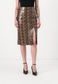 Jupe en simili-cuir serpent marron avec une fente haute, en matériau brillant. Associée à des chaussures à talons noirs.