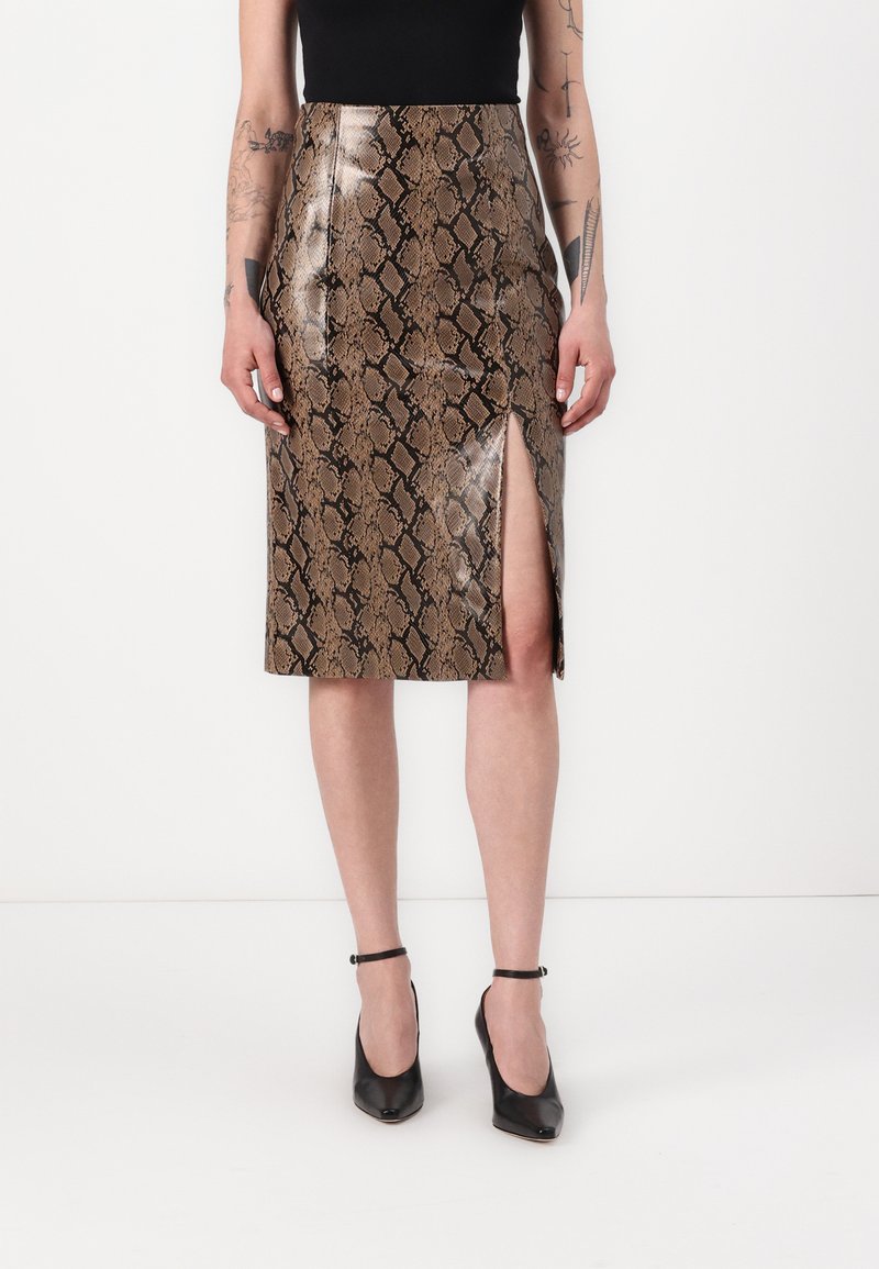 Jupe en simili-cuir serpent marron avec une fente haute, en matériau brillant. Associée à des chaussures à talons noirs.