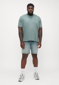 T-shirt di cotone verde chiaro con scollatura rotonda e piccolo logo giallo sul petto, abbinata a pantaloni shorts di denim e scarpe da ginnastica.