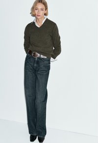 Oliwkowy sweter w serek, zestawiony z wysokim stanem ciemnych dżinsów i czarnych czółenek na szpic. Srebrny pasek podkreśla talię.