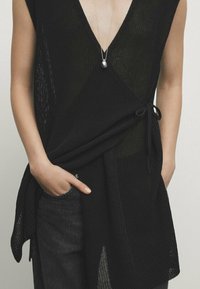 Persoon in een zwarte mouwloze gebreide wikkeltop met een V-hals en een zilveren hangertje, met de handen in de zakken van donkere broek.