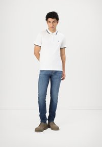 White polo shirt with navy striped collar and small logo, paired with blue denim jeans and grey suede shoes with lace-up design.