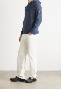 Felpa blu marina con patch logo, pantaloni bianchi a vestibilità rilassata e scarpe nere slip-on con fibbia argentata. Tessuto morbido, design casual.