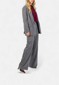 Blazer grigio su misura e pantaloni a gamba larga, abbinati a una maglia bordeaux. Presenta una texture liscia e linee nette. Tacchi scuri e appuntiti.