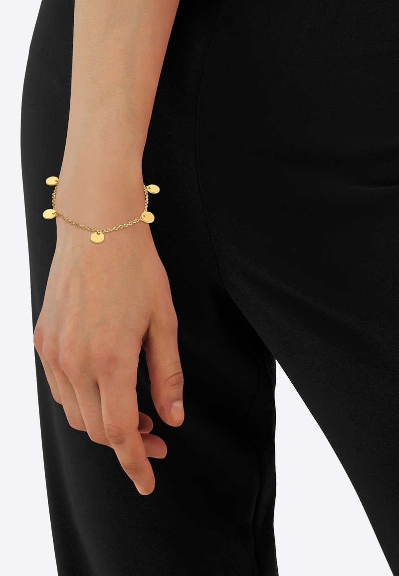 Poignet avec un bracelet chaîne en or comportant cinq petits charms ronds, positionné sur un fond de pantalon noir.