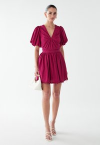 Fuchsia Kleid mit Puffärmeln, tiefem V-Ausschnitt, gerafften Details, elastischem Bund und gewelltem Saum; kombiniert mit einer weißen Handtasche.