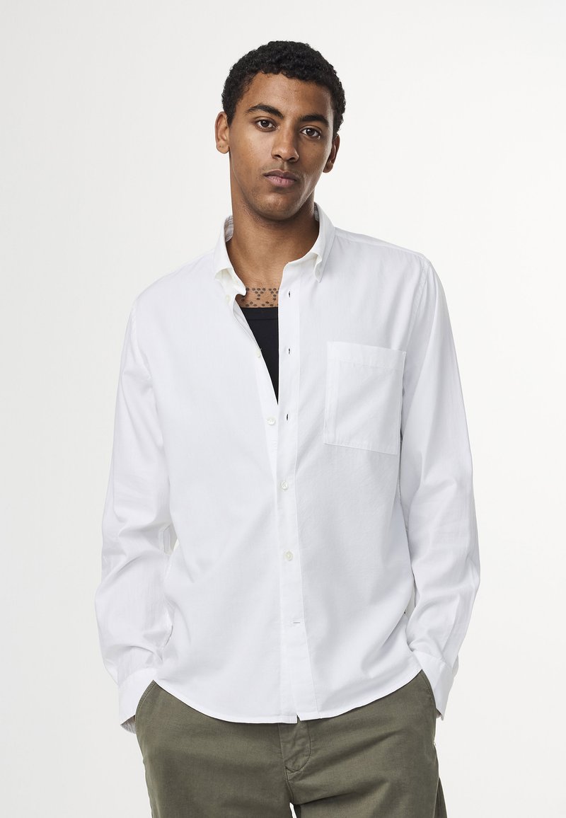 Witte blouse met lange mouwen en knopen, met een kraag, een enkele borstzak en een gladde textuur, gedragen over een zwart T-shirt.