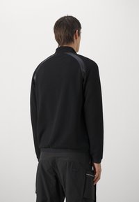 Giacca a pullover nera con corpo in rete testurizzata, accenti neri lucidi sulle spalle e colletto alto. Caratterizzata da una vestibilità rilassata e polsini a coste.
