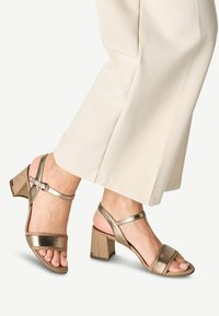 Sandales à talons métalliques bronze avec larges brides à l'avant, bride de cheville et talon carré. Présentent une texture lisse et un design minimaliste.