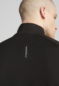 Zwart sportshirt met hoge kraag, een gladde textuur en een reflecterend logo met gestileerde diagonale lijnen op de rug.