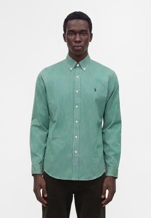Homme portant une chemise à rayures verticales vertes et blanches avec manches longues et un petit logo brodé sur la poitrine.