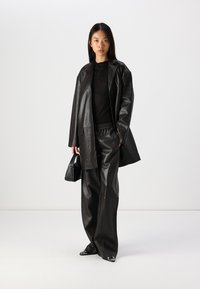 Zwart leren oversized blazer gecombineerd met een aansluitend zwart topje en losse zwarte leren broek. Heeft een gladde textuur en minimale hardware.