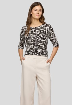 Young woman stands with hands in pockets wearing black and white polka dot top and cream wide-leg pants on plain background.