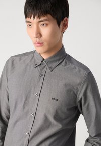 Camisa gris tipo botón con cuello estructurado, mangas largas y bordado de logo en el lado izquierdo del pecho. Presenta botones de metal y una textura suave.