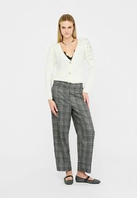 Hvid strikket cardigan med perlebesætning på den ene skulder, kombineret med grå ternede bukser og sorte flade sko. Sat op mod en ensfarvet baggrund.