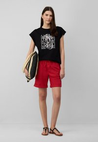 Zwarte katoenen t-shirt met witte grafische tekst, rode opgerolde shorts, zwarte sandalen en een zwart en beige geweven tas. Minimalistisch ontwerp.