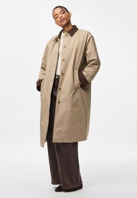 Μπεζ μακρύ παλτό με καφέ γιακά, κλείσιμο με κουμπιά και πλαϊνές τσέπες, συνδυασμένο με σκούρες καφέ φαρδιές παντελόνες και μαύρες μυτερές γόβες.