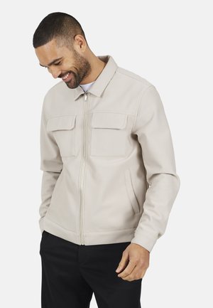 Mężczyzna w beżowej kurtce zapinanej na zamek z kieszeniami na piersi i czarnych spodniach, patrzący w dół i uśmiechający się na tle jasnego, jednolitego tła.