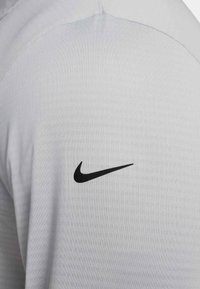 Világosszürke sportpóló, bal vállán fekete Nike jellegzetes pipa logóval, sima, texturált anyagból készült.