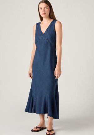 Jeune femme portant une robe midi en denim bleu foncé sans manches avec décolleté en V, ourlet volanté et sandales plates marron, debout sur un fond uni.