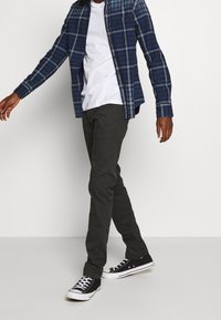 Uomo che cammina indossando sneaker nere, pantaloni scuri, una maglietta bianca e una camicia a quadri blu sbottonata su uno sfondo neutro.