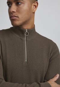 Brown knitted sweater with a high collar, featuring a silver zipper and textured pattern. The fabric appears soft and warm.