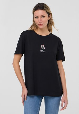 T-shirt in cotone nero con maniche corte, caratterizzata da un design di ricamo floreale in rosa e bianco con il testo "Fiorisci con Grazia" sotto.