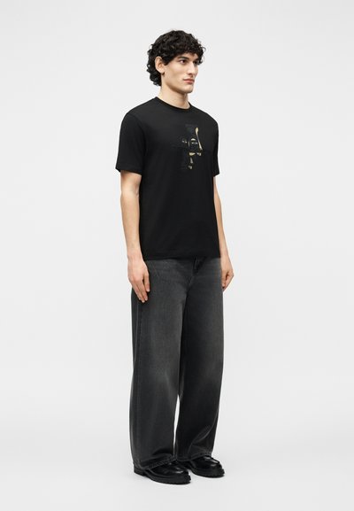 Νέος άνδρας όρθιος, φοράει μαύρο γραφικό t-shirt, μαύρο ξεθωριασμένο τζιν με φαρδιά πόδια και μαύρα παπούτσια, μπροστά σε ένα απλό φωτεινό φόντο.