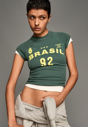 Groen kortemouwengesneden T-shirt met "BRASIL 92" in gele letters; witte onderlaag zichtbaar; gedragen met grijze joggingsbroek om de taille geknoopt.
