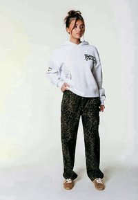 Weißer Hoodie mit schwarzem grafischem Text, kombiniert mit olivgrünen Leo-Print-Hosen. Strukturierter Stoff, lockere Passform und braune Schuhe.