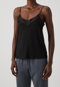 Débardeur noir avec bordure en dentelle, fines bretelles ajustables, tissu lisse et encolure en V. Associé à un pantalon gris ample.