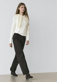 Witte top met lange mouwen, crème trui losjes over de schouders gedragen, donkergroene broek met wijde pijpen, getextureerde bruine enkellaarzen, minimalistische achtergrond.