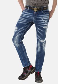Blaue Denim-Jeans mit einer schmalen Passform, ausgefransten Stellen, Waschungen und kontrastierenden Nähten. Getragen mit einem schwarzen Gürtel und Sport Schuhen.