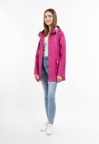 Jachetă waterproof roz cu glugă, dotată cu nasturi tip capse și buzunare. Asociată cu blugi albaștri deschis și adidași albi, pe un fundal simplu.