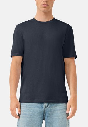 Mężczyzna w prostym ciemnogranatowym krótkim T-shircie z małym logo na piersi i jasnoniebieskich dżinsach, stojący na białym tle.