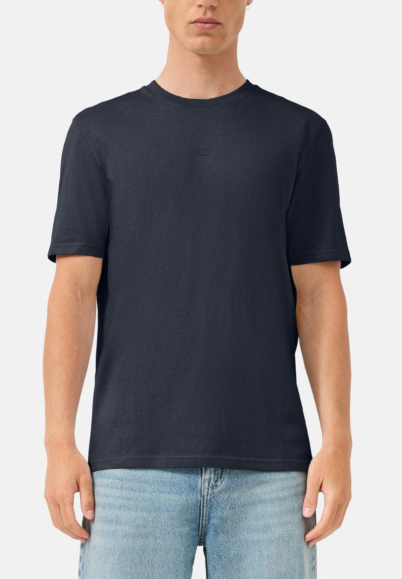 Man draagt een effen donkerblauw (navy) T-shirt met korte mouwen en een klein logo op de borst, en lichtblauwe spijkerbroek, staand tegen een witte achtergrond.