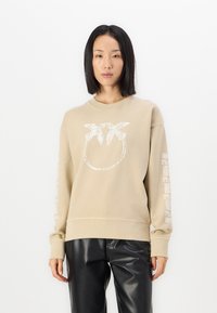Beige pulóver kör alakú grafikával és fehér pálma mintával, hosszú ujjakkal és szövegmintákkal. Fényes fekete nadrággal párosítva.